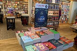 A book table with a display of contemporary romances within a Barnes & Noble bookstore. Most of the books have the “cartoon covers” that are currently popular with publishers. A sign on the display “I’ve Got My Love to Keep Me Warm” and offers a buy one, get one for half price discount.