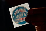 Image of a person holding an “I Voted” sticker. Photo via Unsplash, courtesy of Manny Becerra (@mannyb).