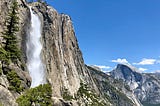 The raging water of Yosemite Falls tumbles over the sheer granite cliff. Half Dome towers in the background.