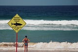 Woman stands in front of the ocean, a sign behind her reads: Shark sighted today, Enter water at own risk