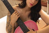 A naked dark-haired female in a cowboy hat, with a pink guitar in front of her.