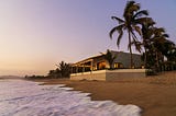 Sunlit modern villa on a palm-lined beach, waves lapping the sand at dusk.