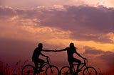 A couple bicycling together in the sunset, the woman is reaching her arm out to the man.