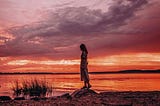A woman stands alone by a lake at sunset, silhouetted against a dramatic sky of purples and reds. Her posture is contemplative, evoking solitude, surrender, and the quiet beauty of letting go.
