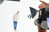 A male model stands in a well-lit studio as the photographer gestures toward him, guiding him into position for a dynamic pose.