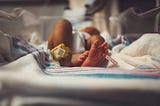 A newborn rests on a hospital bassinet covered with a white blanket featuring pink and blue stripes. The baby’s legs are slightly bent, and the focus is on the tiny feet. One ankle has a yellow monitor band, and the other has a pink hospital ID band. The lighting is soft and warm, highlighting the delicate skin and the peaceful atmosphere of the hospital setting.