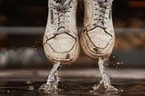 Close-up of beat-up white sneakers jumping up from puddle