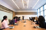 In a conference room, a diverse group of employees with laptops in front of them are facing the whiteboard seeming to listen to another employee speak as she write on the whiteboard.