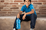 Young boy sitting on a step carrying a water bottle and wearing a pair of sneakers.