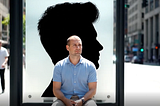 A bald man sitting at a bus stop in front of a large advertisement showing a silhouette of a man with a full head of hair.