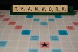 The word “TEAMWORK” is spelled out in Scrabble tiles on top of a game board.