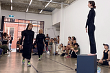 Three dancers stand in a gallery space with white walls. They are all wearing black dresses. One stands upon a grey cube, elevated above the others. An audience watches on.