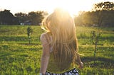 Back of woman with long blond hair, heading to the sun in a big green field