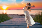 woman wearing long skirt and sleeveless top standing sideways, arms outstretched on a wooden walkway to the beach, sun setting in the distance.