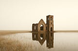 A lone, ruined church stands in calm floodwaters under a pale sky, mirrored by the still surface.