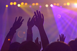 Silhouetten van hoofden en zwaaiende, opgeheven handen in een menigte bij een concert. Paars en geel licht vanaf het podium zijn zichtbaar in de achtergrond.