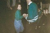 Dad and young daughter dancing in a croweded hall, both wearing green shits on St. Patrick’s Day.