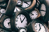 A jumbled pile of wooden analog clocks with a variety of black numerals and dials, set to a wide range of times