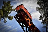 old orange train car suspended on broken track against blue sky with white clouds framed by leafy tree branches