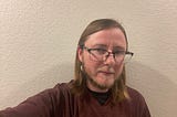 The author is wearing a maroon shirt and is standing in front of a white textured wall. He is wearing black-rimmed glasses, a dangly earring, and has a beard that is gray and brown.