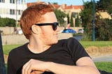 A right-side profile image of the red-headed author wearing black sunglasses and a black t-shirt, and a sunny dark in a park with apartment buildings and road in the background.