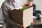 Person wearing a business shirt leaving an office, carrying a box filled with desk contents and a plant.