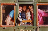 The author’s family of five are inside the first class compartment of an old railway carriage. The author’s mother is holding a bottle of champagne out of the window and everyone is smiling.