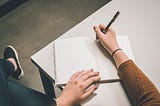 A woman sits at a white desk as she writes her trauma story in a notebook using a black pen.