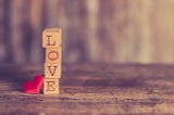 Wooden blocks with letters on them, stand in a stack spelling the word LOVE and beside them a red silk heart, back