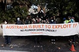 Crowd of people holding banner reading Australia is built to exploit!! No plunder down under #BlockadeAustralia