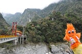 An Orange puppet with googly eyes and a polka-dot vest sticks out over Taiwan’s Taroko Gorge. Behind him an orange and red bridge spans the chasm. Behind this, a pagoda and the Chinese goddess Kuanyin sit among the mountains.