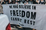 Protesters carrying large white sign with black text “We who believe in Freedom cannon rest”