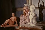 An attractive young woman wearing a beige frilled dress, a white net fascinator, and white lace gloves. She is surrounded by plinths bearing small sculptures, mostly of naked women.