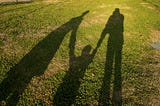 Shadows of a family are shown on a patch of grass. You can see the shadow of a child being held between two shadow parents