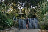 A vacation picture of the protagonist. She casually poses at a blue wooden, surrounded by green ferns and succulents. She wears a dress and sunglasses.