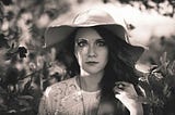 Head shot of a dark-haired woman among trees in a floppy hat with wide-eyed, eerily angry expression.