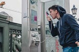 This is an image of a white guy in a jeans and a dark blue hoodie who is screaming into the receiver of a payphone and gesturing angrily.