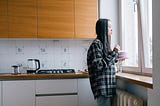 A dark-haired woman stands alone in her kitchen, holding a bowl, and looking out the window. A cooktop and coffee pot are visible behind her, and a radiator is under the windowsill through which she looks at the world.