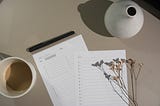 Note pages pre-formatted for checklists of different lengths. Remnants of a light coffee to the left, dried flowers and a vase to the right. The pen rests, capped, across the top of the pages.