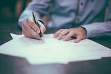 A man’s hand holds a pen. He is signing some documents that are on a table.