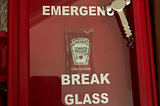 Picture of gag gift featuring a hammer and a packet of ketchup behind glass with “emergency break glass” written on it.