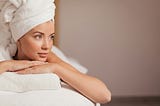 Image shows a woman lying on a massage table. Her hair is pulled up into a towel on top of her head and she is lying on her stomach, arms supporting her chin. Her eyes are open and she looks relaxed.
