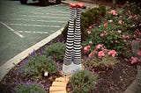 Image shows a pair of legs, sticking up out of the ground in a garden, wearing black and white striped stockings and sparkly ruby red shoes, with a yellow brick road leading to the legs.