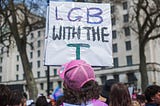 Building in the background. Protest sign held by person with brown curly shoulder length hair, wearing a pink snap back. The Sign is a white rectangle that says ‘L G B with the T’.