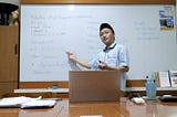 Munip Utama stands before a whiteboard, gesturing. A laptop sits open on the table in front of him.