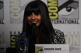 Jameela sits in front of a black and white background, smiling into mic, wearing black and white blouse