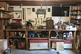 A cluttered work bench with projects in various states of completion. Tools are on the shelves and a radio, TV, and more tools are hung on the wall behind it.