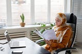 A woman in business attire with a hijab covering her hair in a modern and stylish office sat in front of a computer reading a budget report.