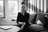 Portrait of Executive Life Coach Signe Jungløw in black and white sitting in a sofa bathed in sunlight with a black kitten on her side and a macbook air on the table in front of her. She is wearing all black.