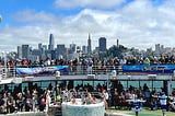 Image of a crowded group of guests and crew dancing and celebrating on the pool deck (level 15) and upper deck (level 16) of the Ruby Princess cruise ship with the San Francisco skyline in the background below a bright blue sky and fluffy white clouds. Two large banners hanging from the guardrails read “Sail Away” party.